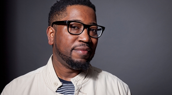a poet with brown skin and hair, light beard and dark framed glasses, blue t-shirt under a buttoned white shirt