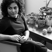 a poet depicted in b&w seated looking over her shoulder, hands joined over a dark dress, legs crossed, dark curly hair cut above the shoulder; behind her an assemblage of vases with plants and other vessels