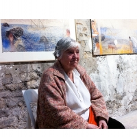 a woman, short grey hair combed back, sits watching before her, in front of a stone wall with colorful paintings, mythic scenes; she's wearing a pink and grey sweater, a white shirt, and holding a bright red handbag in her lap