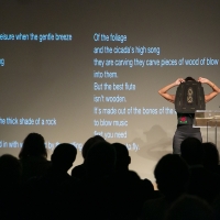woman holding up a masklike fiber structure before her face, elbows akimbo, onstage before an audience, text of a poem projected on wall at her back