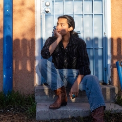 a poet-musician partly in shadow with long straight dark hair, seated on a doorstep, light blue painted door behind them
