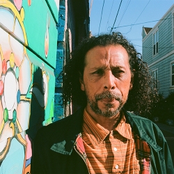 poet in orange striped shirt, green jacket, long brown hair and cropped beard, in Clarion Alley with murals at their back