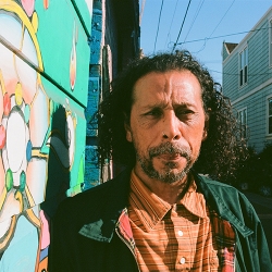 poet in orange striped shirt, green jacket, long brown hair and cropped beard, in Clarion Alley with murals at their back