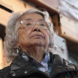 woman in silver hair, light glasses, blue scarf and dark coat gazes upward before a wall of stacked suitcases