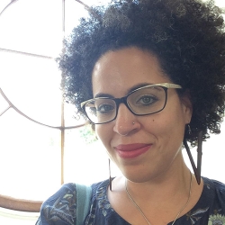 poet with thick curly hair, bold glasses, red lipstick and a collarless dark top, before a window with a circular motif, light streaming thru