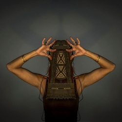 a poet/performer with a leathery text mask before their face, arms akimbo, hands raised to their head, fingers spread, wearing a dark trailing garment