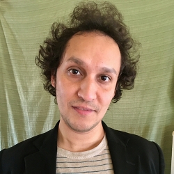 a young man with direct sober gaze looks directly outward—he has dark curly hair, and has on a dark sportscoat over a striped t-shit, while behind him olive colored drapery falls