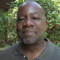 dark brown calmly smiling face, light gray beard cropped short, gray shirt open at collar, red-berried green bush behind