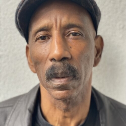 poet with a broad brown mustache, slender face and sharp direct gaze, dark snapbrim cap, grey jacket and dark shirt