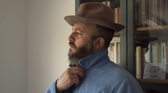 in a sharp brown fedora and blue denim jacket, a man looks toward the window, offscreen, his reflection behind him in a filled glass bookcase