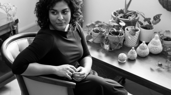 a poet depicted in b&w seated looking over her shoulder, hands joined over a dark dress, legs crossed, dark curly hair cut above the shoulder; behind her an assemblage of vases with plants and other vessels