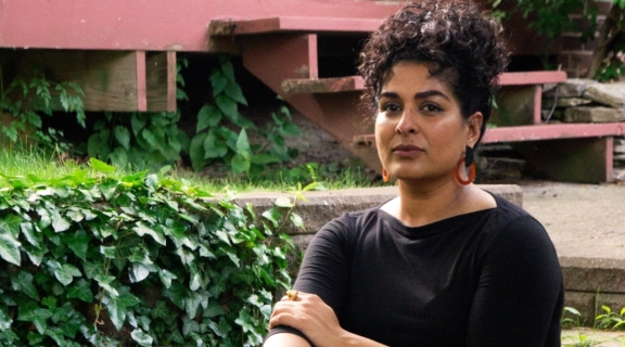 a poet in dark dress, one hand on opposite arm, dark hair piled up, seated on steps before a green garden and deep rose colored house