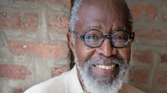 a poet with brown skin, white hair and wide smile, wearing round-rimmed glasses and a white suit, before a brick and mortar wall