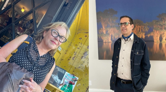 two poets, one in a grey dress dark glasses and silver hair before a brightly painted doorway; one in a dark suit, dark glasses dark hair + white shirt standing before a blue + gold painting on the wall