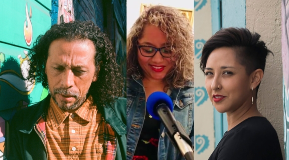 3 poets-one in striped orange shirt + green jacket, long hair + cropped beard; one in curly long hair, jean jacket, dark glasses + red lipstick; one in cropped dark hair + dark shirt before pale blue + pink mural