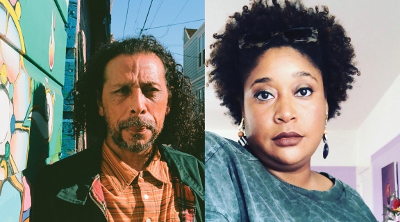 2 poets, one in orange plaid shirt, green jacket, long brown hair and cropped beard, in Clarion Alley; second with blue collarless shirt, curley dark hair + dark mauve lipstick, multicolored flowers in right corner