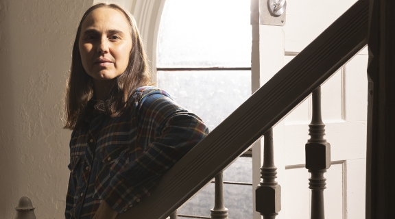 a poet with long straight hair looks into the distance toward the camera, standing with arm leaning onto a stair banister, sunlight spilling from an ovoid window behind