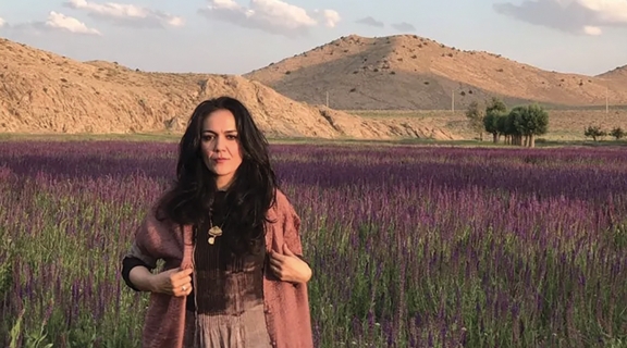 a woman with long dark hair, a long reddish jacket and light brown skirt, before a field of blooming heather, green and brown foothills in the background