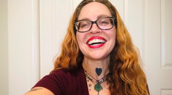 woman with broad smile, red lipstick, dark rimmed glasses, blonde-red long hair, a floral necklace tattoo, open necked purple shirt