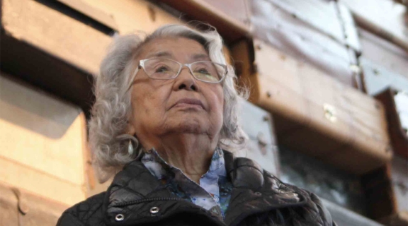 woman looking upward, silver hair and light rimmed glasses, standing before a wall of suitcases