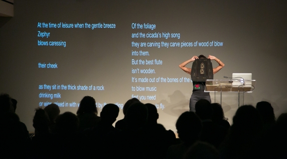woman holding up a masklike fiber structure before her face, elbows akimbo, onstage before an audience, text of a poem projected on wall at her back