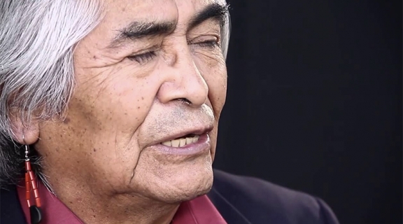 A close-up image of poet Simon Ortiz, from an angle to his right. He has straight grey hair, a red beaded earring, a burgundy shirt collar and dark jacket.
